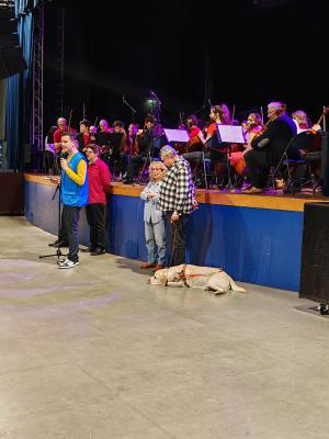Photo d'une scène avec un orchestre et un homme qui parle au micro. Deux personnes représentants l'association sont à ses côtés avec un chien guide.